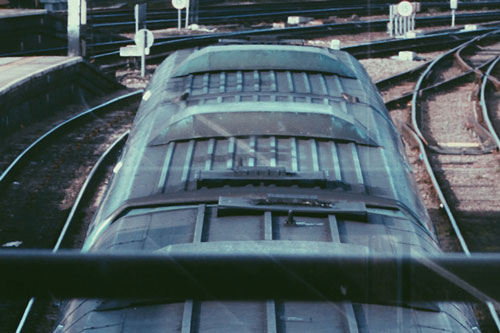 A passenger train seen from above, showing the curved grey roof and adjacent platform and tracks at the station.