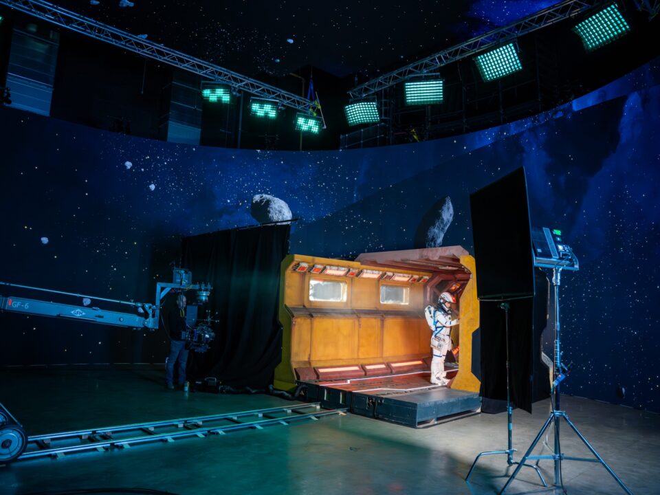 Film crew recording an actor in a space suit against a large curved LED screen displaying an outer-space scene with stars and asteroids.