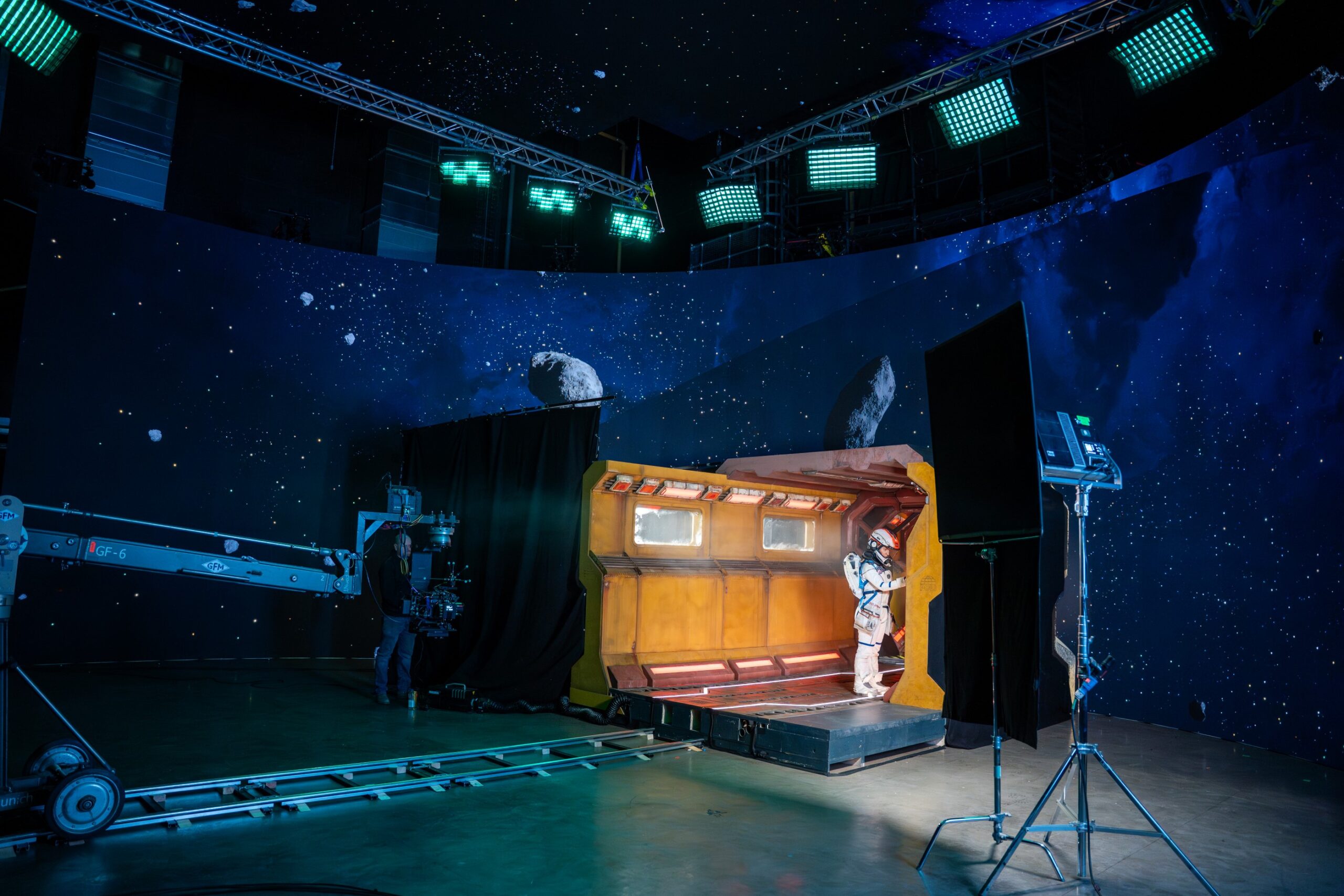 Film crew recording an actor in a space suit against a large curved LED screen displaying an outer-space scene with stars and asteroids.