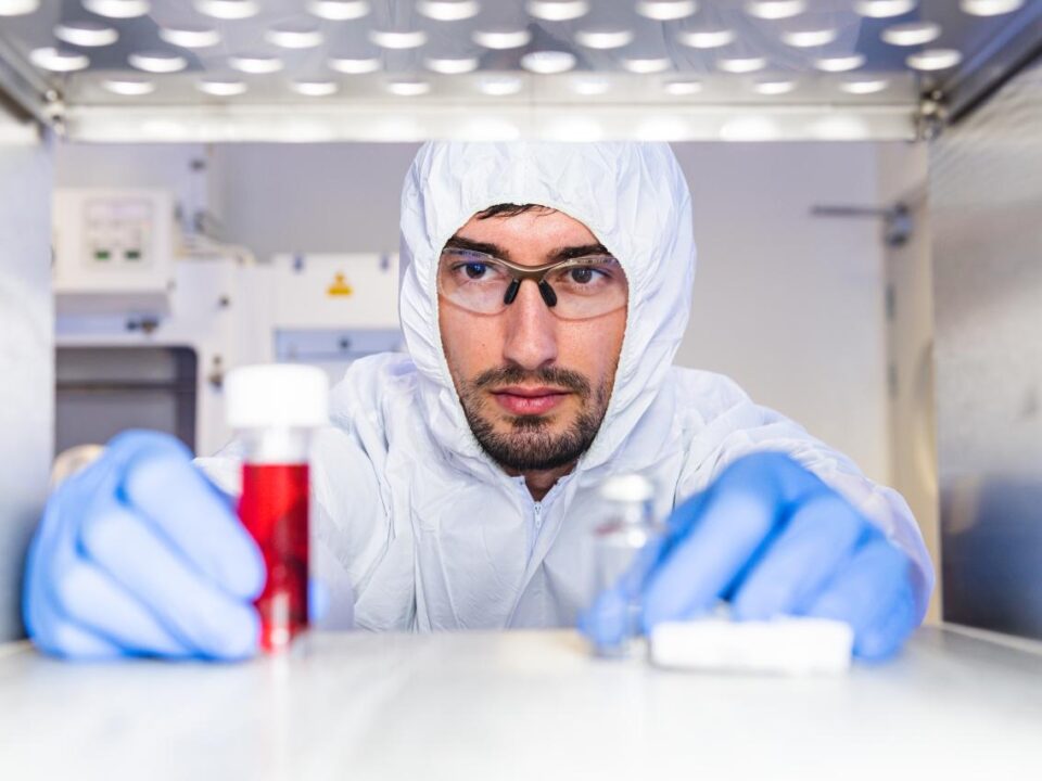 Scientist in protective lab clothing and gloves examining vials, including one containing red liquid, inside a sterile laboratory environment.