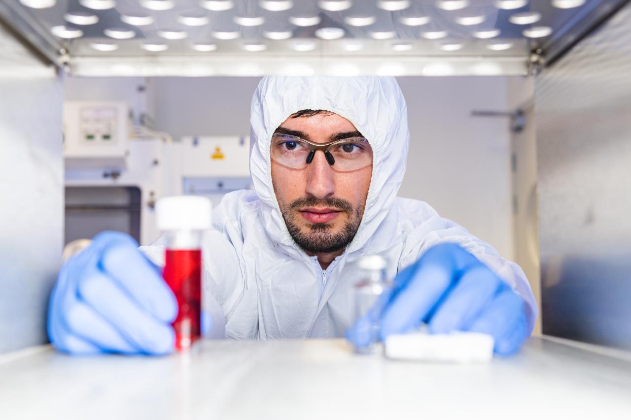 Scientist in protective lab clothing and gloves examining vials, including one containing red liquid, inside a sterile laboratory environment.