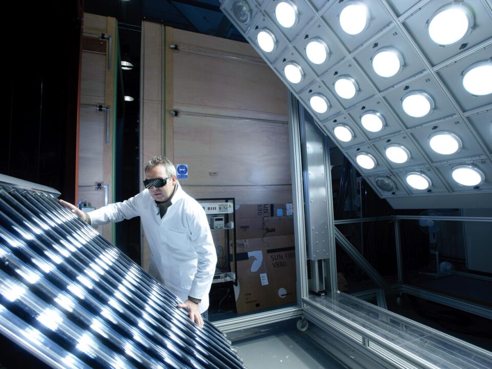 Researcher in a white lab coat and protective glasses operating solar energy testing equipment under an array of bright simulated sunlight lamps.
