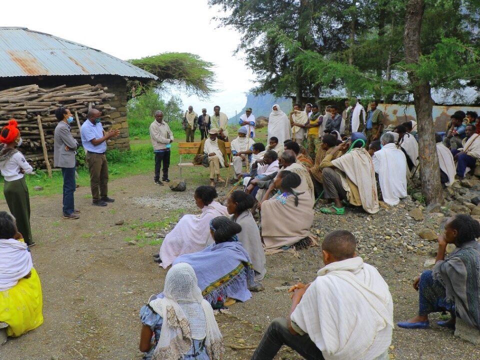 A community health education session is taking place outdoors in a rural setting, with local residents gathered to listen to facilitators speaking.