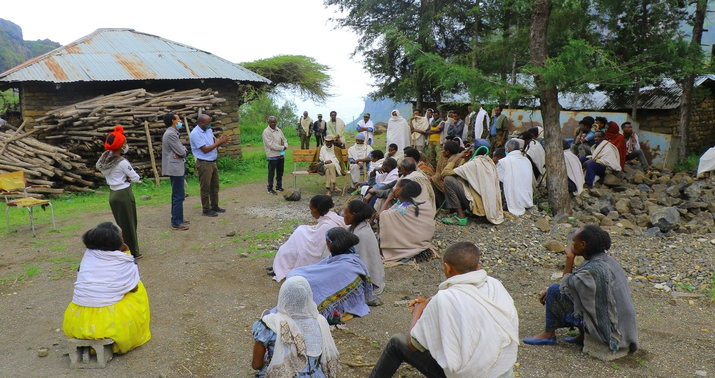 A community health education session is taking place outdoors in a rural setting, with local residents gathered to listen to facilitators speaking.