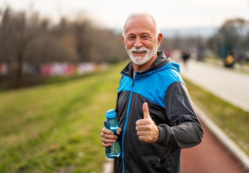An older man in sportswear smiles and gives a thumbs-up while holding a water bottle outdoors.