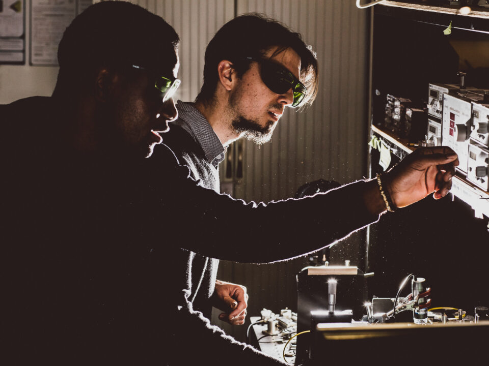 Two researchers wearing safety glasses work together in a dimly lit laboratory, adjusting equipment on a workbench illuminated by focused light.